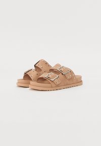 Le ciabatte in suede color tan presentano due ampie cinghie, fibbie in argento e borchie decorative. La soletta è piatta con una suola in gomma testurizzata.