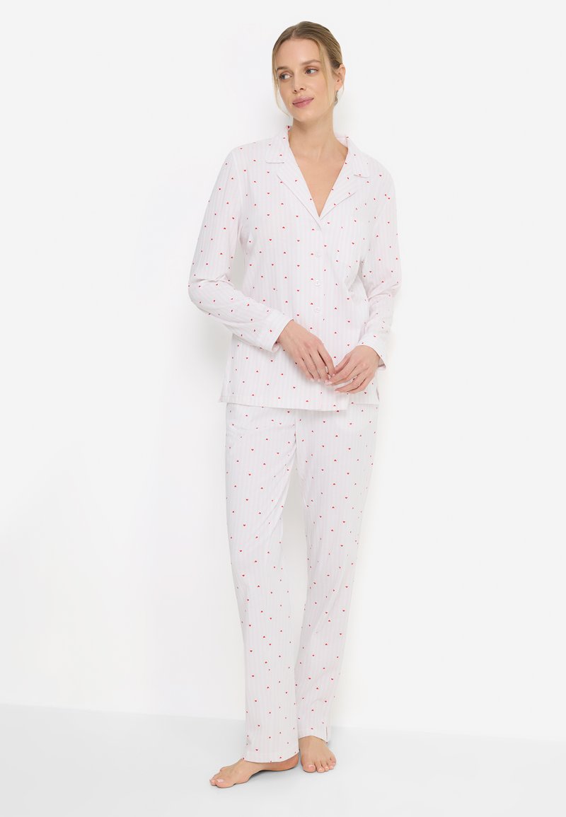 Λευκή πιτζάμα με κόκκινο μοτίβο καρδιάς, που περιλαμβάνει μπλούζα με γιακά και κουμπιά, καθώς και μακριά παντελόνια με ίσια εφαρμογή και ελαφρύ ύφασμα.