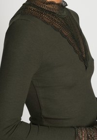 Långärmad topp i mörkgrön ribbad tyg, med en hög spetskrage med intrikata detaljer och en figursydd silhuett.