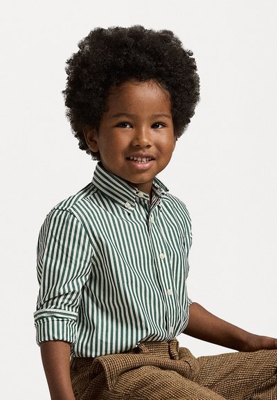 Ραγισμένο πουκάμισο σε πράσινο και λευκό, με γιακά και κυλιόμενα μανίκια, σε συνδυασμό με υφή καφέ παντελόνι. Παιδί καθιστό, χαμογελαστό.
