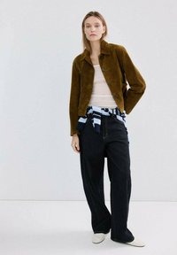 Mujer con chaqueta marrón con botones, camiseta blanca, vaqueros negros de pierna ancha y un suéter a rayas atado a la cintura, posando frente a un fondo liso.