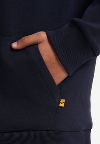 Sweatshirt blu navy con tasca a marsupio, polsini a costine e una piccola etichetta gialla sulla cucitura. Tessuto morbido e texturizzato.