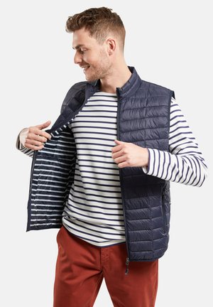 Gilet matelassé marine avec col haut et fermeture éclair, porté sur une chemise à manches longues blanche et marine à rayures, avec des poches latérales.
