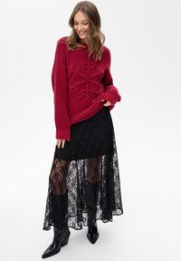 Czerwony dzianinowy sweter z teksturowanym wzorem śnieżynki, zestawiony z długą czarną koronkową spódnicą maxi i czarnymi botkami. Gładkie, warstwowe tekstury.