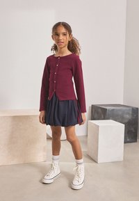 Long-sleeve burgundy ribbed top with buttons, paired with a navy puff skirt featuring red side stripes and white high-top sneakers. Smooth texture.