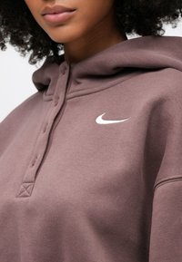 Κοντινό πλάνο ενός ατόμου που φοράει μωβ φούτερ με κουμπωτό γιακά και λευκό λογότυπο Nike στο στήθος.