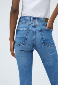 Persona che indossa dei jeans blu e una camicia bianca, con la mano destra nella tasca posteriore e la mano sinistra rilassata lungo il fianco.