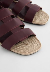 Les sandales à enfiler en cuir bordeaux présentent de larges brides tissées sur une semelle intérieure en corde naturelle avec un design plat et texturé.