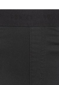 Gros plan sur la ceinture en tissu noir avec le logo "HUGO" embossé en répétition et la couture verticale visible sur le vêtement.