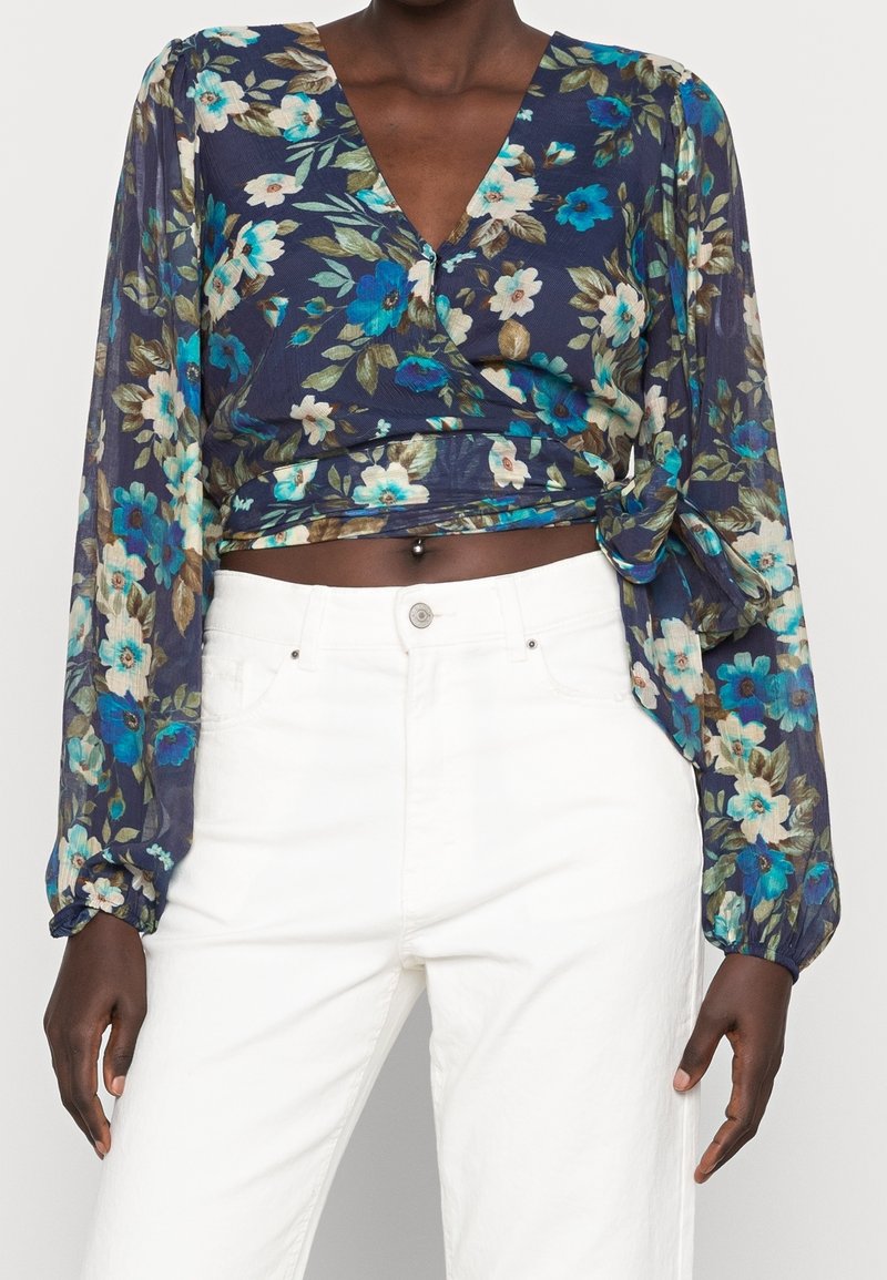 Blouse portefeuille fleurie bleu marine avec un décolleté en V profond, ornée de fleurs bleues et blanches, manches longues bouffantes et détail noué à la taille. Assortie à un pantalon blanc.