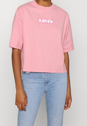 Persona vistiendo una camiseta corta rosa de Levi's con mangas cortas y jeans azul claro, de pie contra un fondo liso.