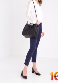 Sort pose læder håndtaske med et struktureret design, snørre lukning, flad bund og dobbelte bæreremmer. Modellen har en hvid bluse og navy bukser på.