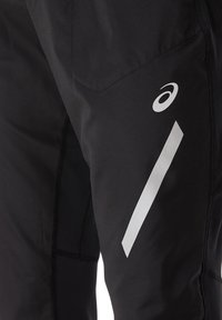 Pantalon de sport noir avec logo blanc réfléchissant ASICS et bande diagonale sur la cuisse, montrant la texture du tissu et les détails des coutures.