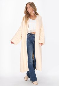 Langer, cremfarbener Strickcardigan mit strukturierten, voluminösen Ärmeln. Getragen über einem weißen kurzen Top und blauen Schlagjeans. Weicher, kuscheliger Stoff.