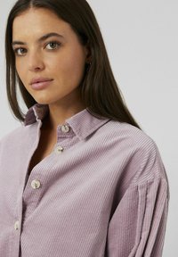 Chemise en velours côtelé violet clair avec col, dotée de fermetures à boutons et d'une texture ribbée. Design ajusté avec des manches légèrement froncées.