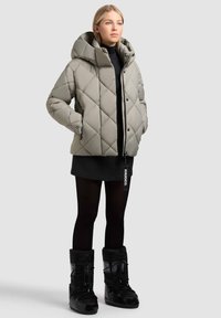 Lys grå pufferjakke med diamant quiltet mønster, stor hætte og lynlås lukning. Båret over en sort top og sorte tights med sorte snestøvler.
