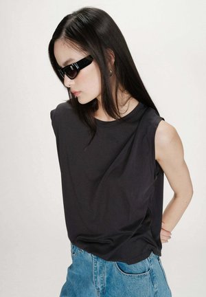 Femme aux longs cheveux foncés portant un haut noir sans manches, un jean bleu et des lunettes de soleil noires, debout les mains derrière le dos sur un fond blanc.