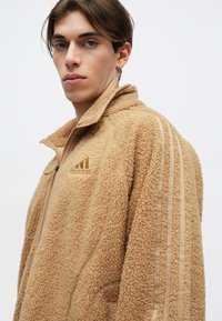 Fiatal személy bézs textúrájú Adidas zippes dzsekit visel, három csíkkal a kézfején és egy kis karikafülbevalóval, egyszínű háttér előtt.