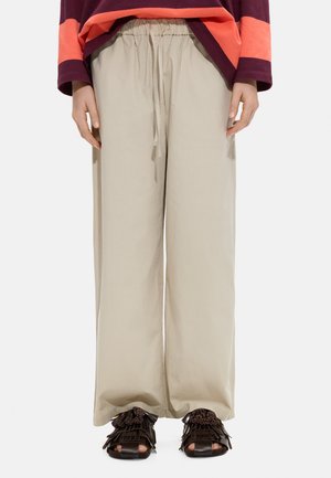 Beżowe spodnie o szerokich nogawkach z paskiem ze sznurkiem w talii, noszone z czarnymi sandałami z otwartym czubkiem i warstwowymi frędzlami oraz pasiastą bluzką z długim rękawem.