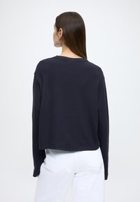 Tmavomodré tričko s dlhými rukávmi, voľného strihu, žíhanou textúrou a okrúhlym výstrihom. Bez viditeľných vzorov alebo kovových doplnkov. Pohľad zozadu ukazuje jednoduchý dizajn.