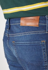 Jean en denim bleu foncé avec une texture lisse, des coutures jaunes contrastantes et un patch en cuir marron portant l'étiquette "JACK & JONES".