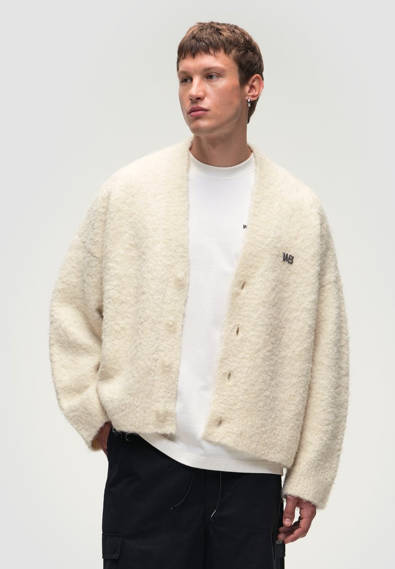 Cardigan di colore crema, soffice e dal taglio rilassato, con colletto a scialle e sei bottoni frontali. Accentuato da un logo.