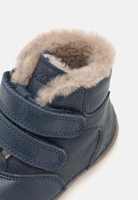 Botă din piele albastră închisă cu două curele Velcro, blăniță moale de culoare bej în jurul gâtului, și o talpă texturată pentru aderență.