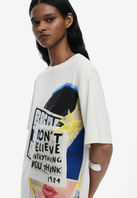 Yliantennettu valkoinen puuvillat-paita, jossa on graafinen printti, jossa on kasvokuva, tekstiä ja värikkäitä muotoja, mukaan lukien sininen tähti ja sanomalehtileikkeitä.