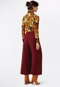 Haut en turtleneck floral à manches longues et ajusté dans des tons chauds, associé à un pantalon large taille haute bourgogne et des bottes à talons beiges.