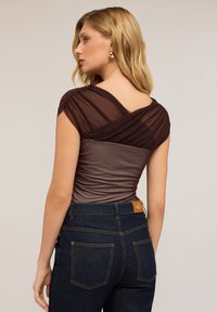 Donna con capelli biondi, che indossa orecchini a cerchio dorati, un top trasparente marrone a incrocio e jeans in denim blu scuro con tasche posteriori visibili.