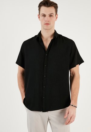 Camisa negra de manga corta con un frente de botones, cuello y una tela texturizada. Se lleva con pantalones de color claro para crear contraste.