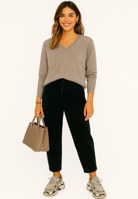 Beige v-hals trui, zwarte high-waisted jeans, grijze sneakers met donkere accenten, en een beige handtas. Glad textuur overal.