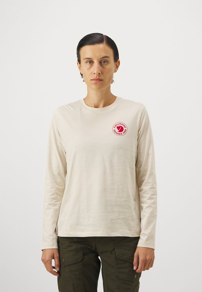 Beige pikkade varrukatega särk pehmest kangast, millel on vasakul rinnal ümmargune punane logoetikett. Lihtne disain, ümmarguse kaelusega.