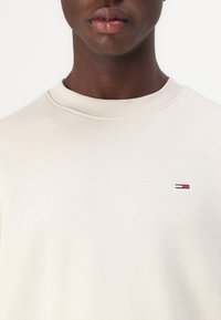 Lys beige genser med ribbestrikket rund hals, prydet med en liten brodert logo i rødt, hvitt og blått på venstre bryst. Myk tekstur.