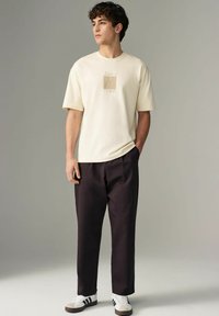 Cremfarbendes T-Shirt mit kurzen Ärmeln und geprägtem Picasso-Design, kombiniert mit dunklen Hosen und weißen Schuhen mit schwarzen Streifen.