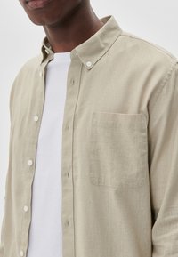 Light beige button-up shirt made of textured fabric, featuring a collar, white buttons, and a chest pocket. Worn over a white T-shirt.