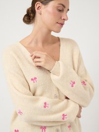 Κρεμ ζακέτα με ριμπ υφή, διακοσμημένη με ροζ κέντημα φιόγκου και στρογγυλά κουμπιά στη μπροστινή πλευρά.