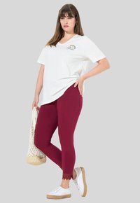 Camiseta blanca de cuello en V con un gráfico que dice "Bloomed & Pure", combinada con leggings burdeos con encaje en el tobillo y zapatillas blancas.