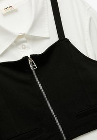 Top corto nero con chiusura a zip, realizzato in tessuto morbido. Presenta spalline regolabili e due tasche frontali. Abbinato a una camicia bianca con colletto.