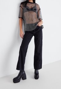 Mujer vestida con una blusa de malla negra transparente sobre un sujetador negro, pantalones negros de pierna ancha de cintura alta y botas de plataforma negras hasta el tobillo, apoyada contra una pared blanca.