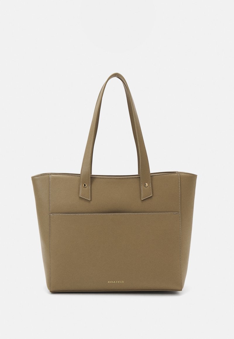 Sac fourre-tout rectangulaire de couleur taupe avec longues anses et détails de couture à l'avant, avec petits lettrages dorés du nom de la marque près du bas.