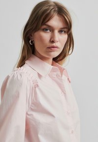 Šviesiai rožinė marškinė su mygtukais, raukšlėmis pečių siūlėse, apykakle ir švelnia audinio tekstūra. Subtili blizgesys su apvaliais mygtukais.