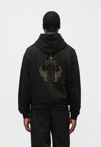 Čierna mikina s kapucňou s veľkým zlatým grafickým motívom na chrbte, zobrazujúca psie hlavu a kvetinové prvky, navrhnutá s voľným strihom a žebrovanými manžetami.