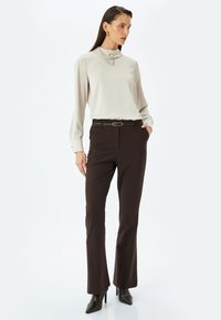Blusa de color beige claro con un collar alto y un botón decorativo, combinada con pantalones rectos marrón oscuro y zapatos de tacón negros.