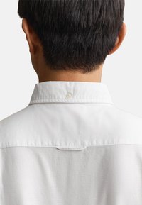 Vue de dos d'une personne aux cheveux courts et foncés portant une chemise blanche à col sur un fond uni.