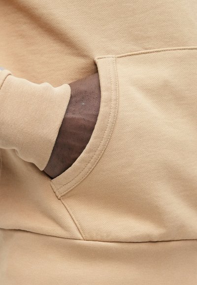 Ύφασμα μπεζ φούτερ με υφή, διαθέτει μπροστινή τσέπη και ριμπ μανσέτες. Κοντινή άποψη της λεπτομέρειας της τσέπης.