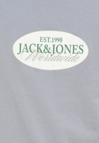 Ανοιχτό γκρι ύφασμα με λευκή οβάλ ετικέτα λογότυπου που εμφανίζει το κείμενο "EST. 1990 JACK & JONES Worldwide" σε πράσινο  και γκρι χρώμα.