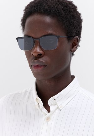 Giovane con capelli corti e ricci che indossa occhiali da sole quadrati scuri e una camicia bianca con sottili righe verticali.
