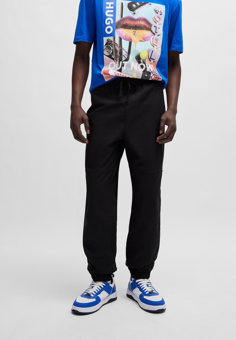 Sorte joggingbukser med elastik i taljen, tilspidsede manchetter og sidesømme. Bruges med en blå grafisk t-shirt og blå/hvide sportssko.