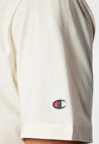 Cremfarbene Baumwolle Kurzarm-T-Shirt mit Rundhalsausschnitt und einem gestickten Logo auf dem linken Ärmel in Schwarz, Rot und Weiß.